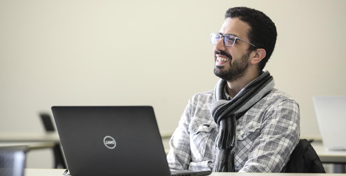 一个学生坐在笔记本电脑后面，对着镜头外的人微笑. 这个学生戴着眼镜，穿着格子衬衫，围着围巾.