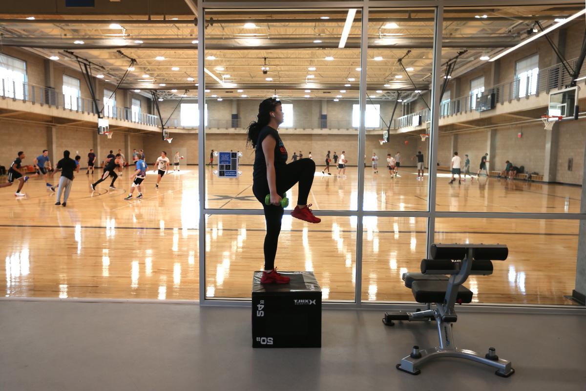 一个学生在玻璃墙的房间里举重.你可以看到篮球场在墙的另一边, 一群人在哪里一起打篮球.
